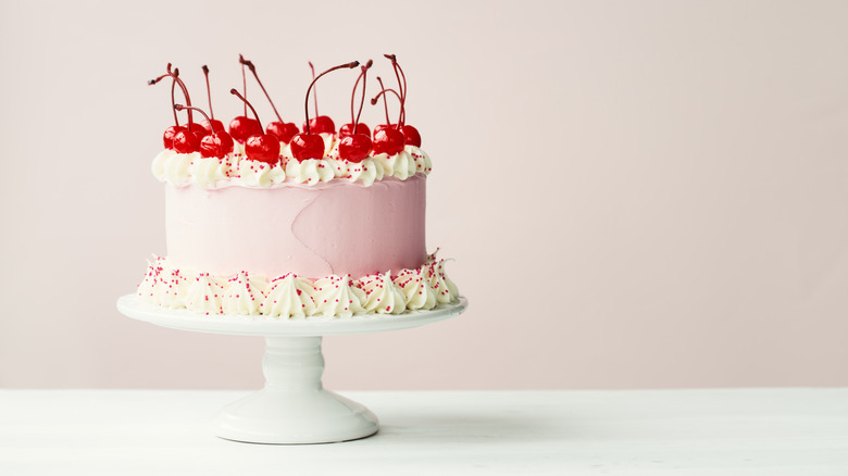 Cherry chip cake in pink frosting topped with cherries on cake stand.