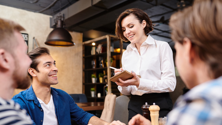 Smiling pretty waitress with short hair wearing white blouse standing at table with guests and making notes in sketchpad while taking order in restaurant, guys flirting with her