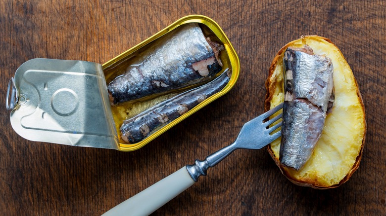 Open tin can of sardine fish with fork and baked half potato