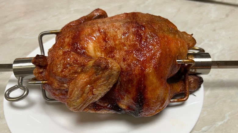 Freshly baked rotisserie chicken still on the skewer sitting on round white plate on kitchen bench