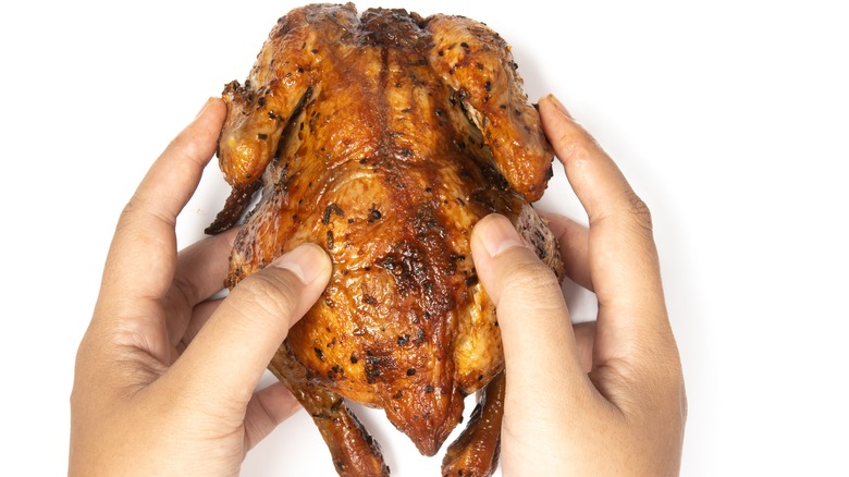 Hand holding a whole delicious roasted chicken seasoned with herbs on white background