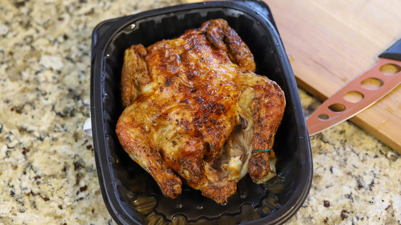 An organic rotisserie chicken in a black plastic container on a kitchen counter next to a wooden board and sharp knife