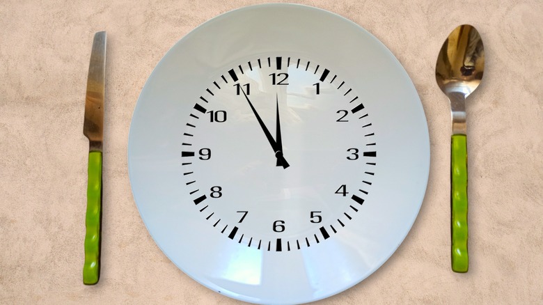 A top-down view of a clock set at five to 12, drawn on a white plate with green-handled cutlery on a beige background