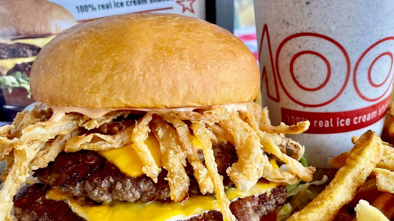 Burger on a bun with onion strings, cheese, lettuce, and tomato beside fries and a milkshake