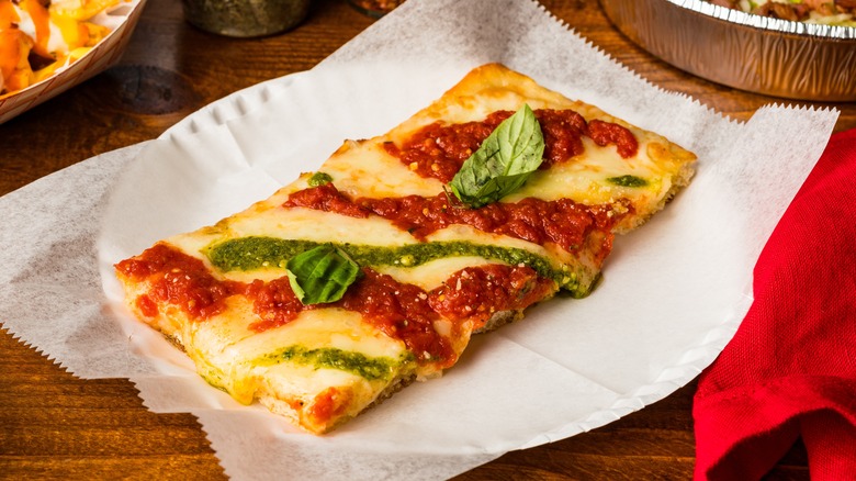Slice of Grandma pizza on a paper plate with pesto and tomato sauce