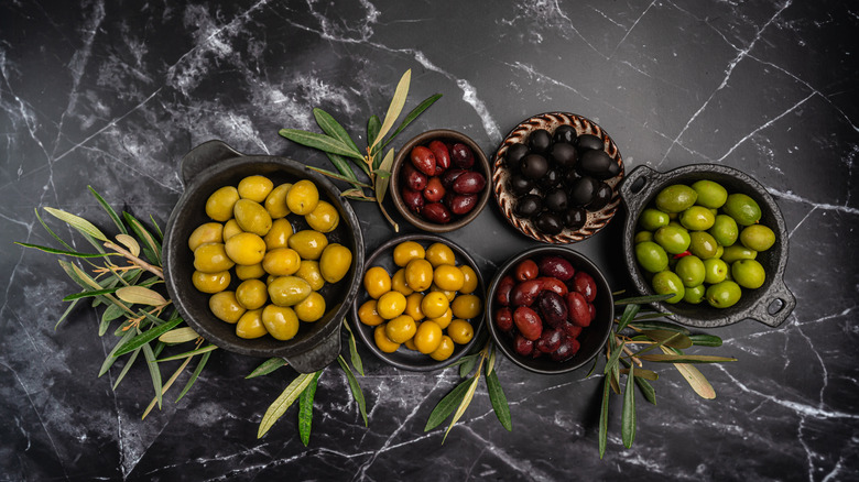 Green and black olives in a bowls and olive oil on dark background top view. Olive oil and variety of green and black olives in bowls with olive leaves all around taken from above