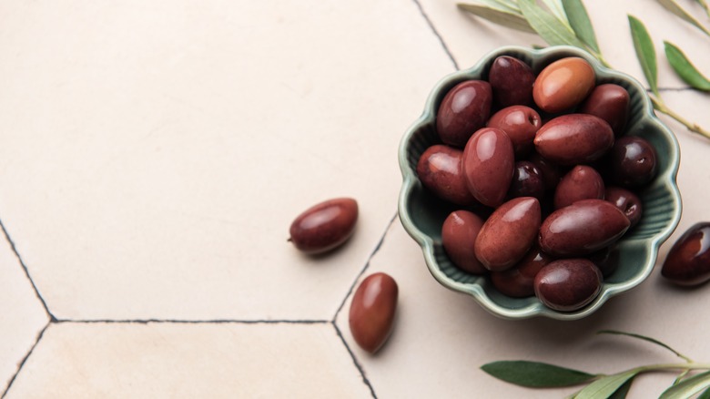 Kalamata olives and olive branches are arranged in a green bowl on a tiled surface