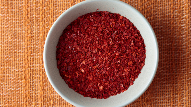 A white bowl of gochugaru red chili flakes on an orange woven mat