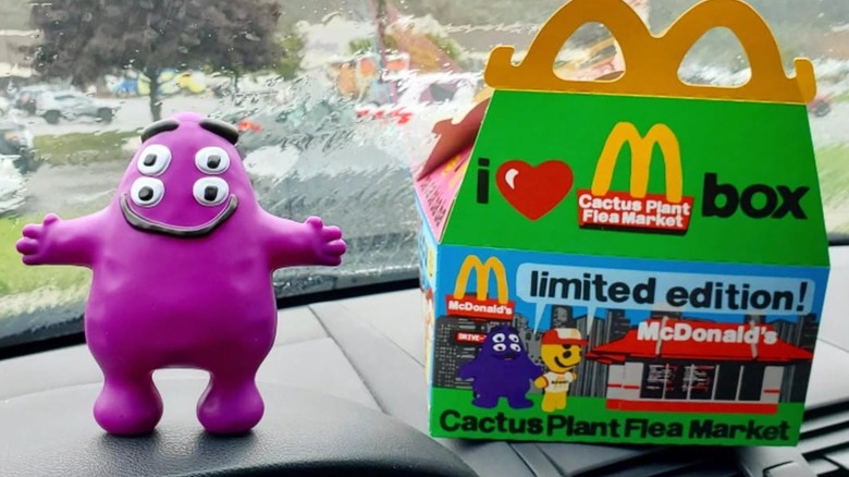 A Cactus Plant Flea Market Happy Meal box with a Grimace toy on a car dashboard