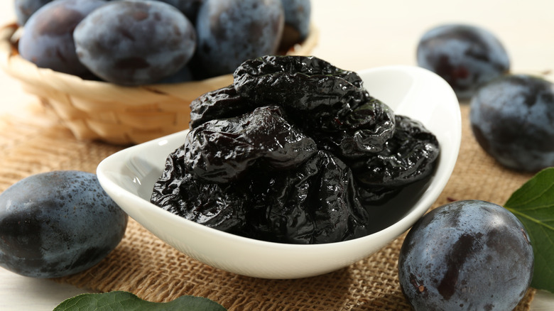 Dried prunes in a white bowl surrounded by fresh plums