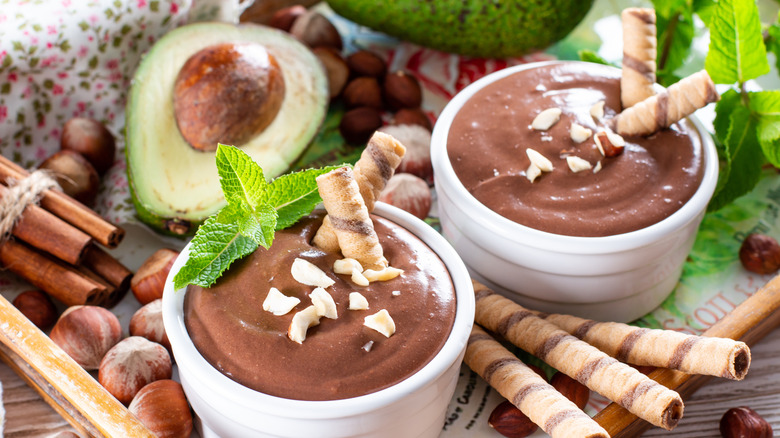 Two cups of chocolate pudding surrounded by avocado and praline sticks.