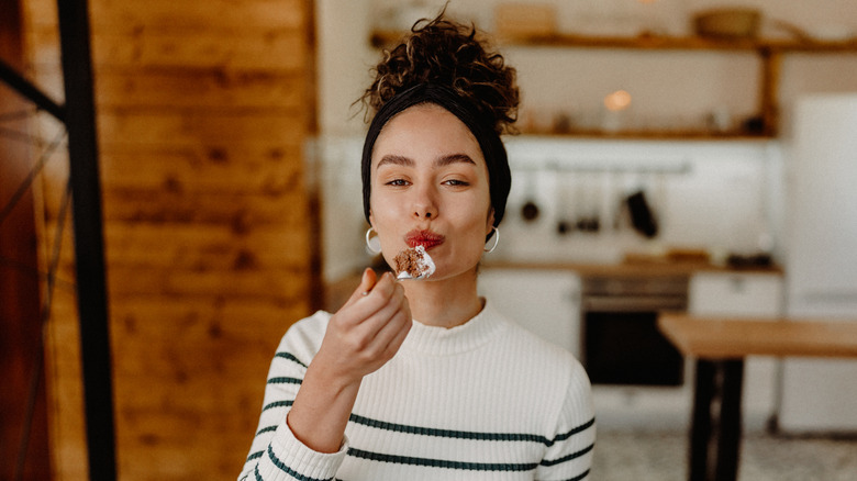 Young woman standing in kitchen eating slice of cake.