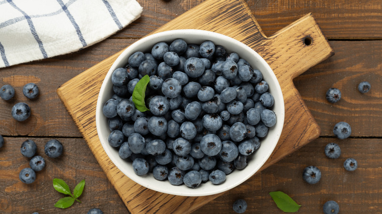 A white bowl of fresh blueberries on a wooden cutting board