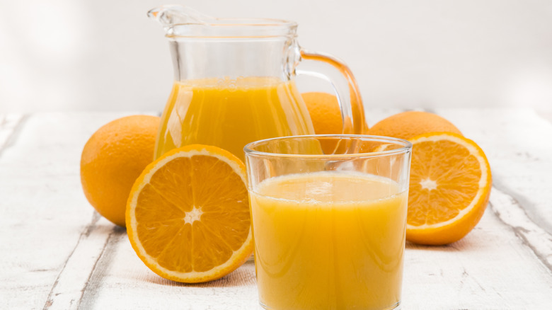 pitcher of orange juice and glass of orange juice on a light background with sliced oranges surrounding them