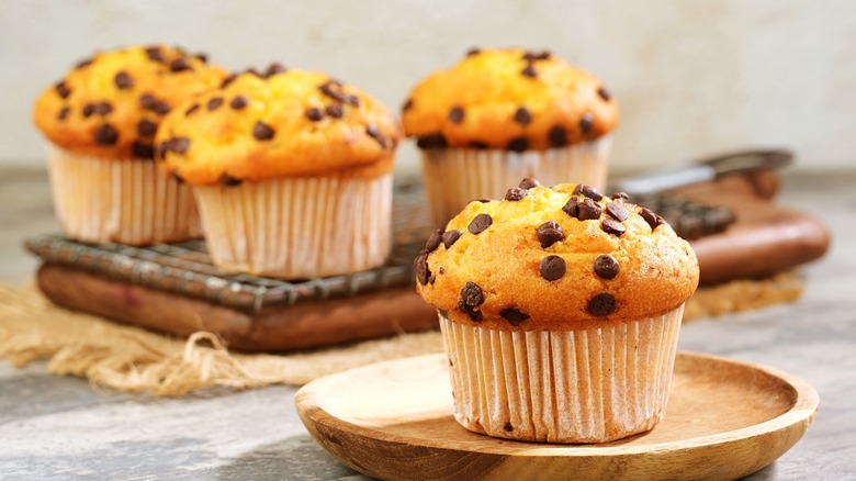 A close-up of a chocolate chip muffin on a wooden plate, with more muffins on a wooden cutting board in the background