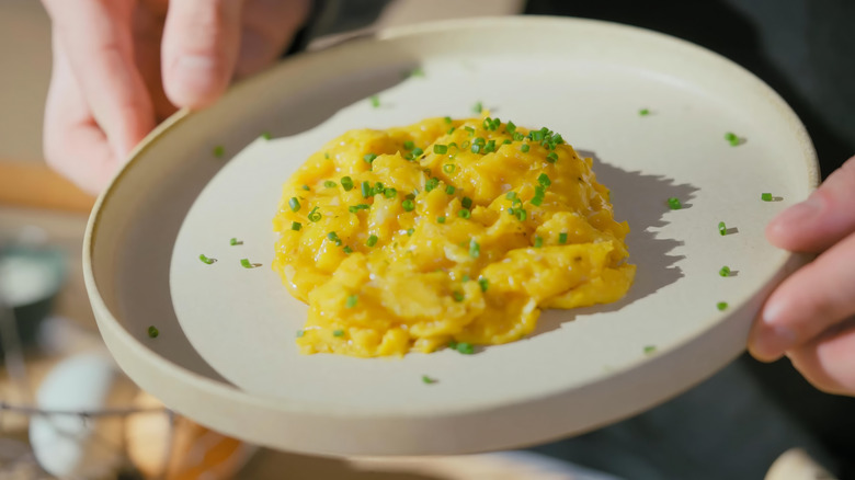 hands holding white plate of creamy scrambled eggs topped with chives.