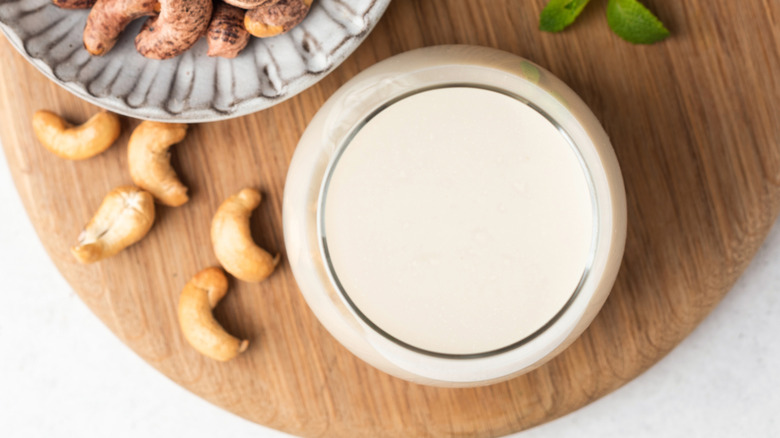 Top-down view of a glass of cashew milk on a wooden board, next to some loose nuts and some on a plate