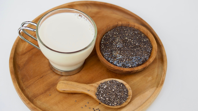 A small glass of milk next to a wooden spoon and bowl full of chia seeds on a wooden board on a white background