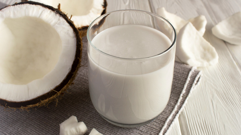 Close up of a glass of ccoconut milk sitting on a pale grey cloth, next to two open halves and several pieces of coconut on a white wooden surface