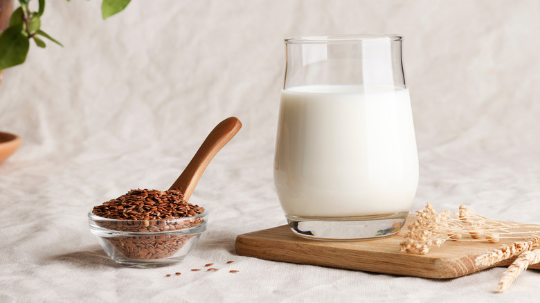 flax seed milk in glass and dried flak stems on a wooden board, next to a glass bowl of flax seeds with a wooden spoon in it