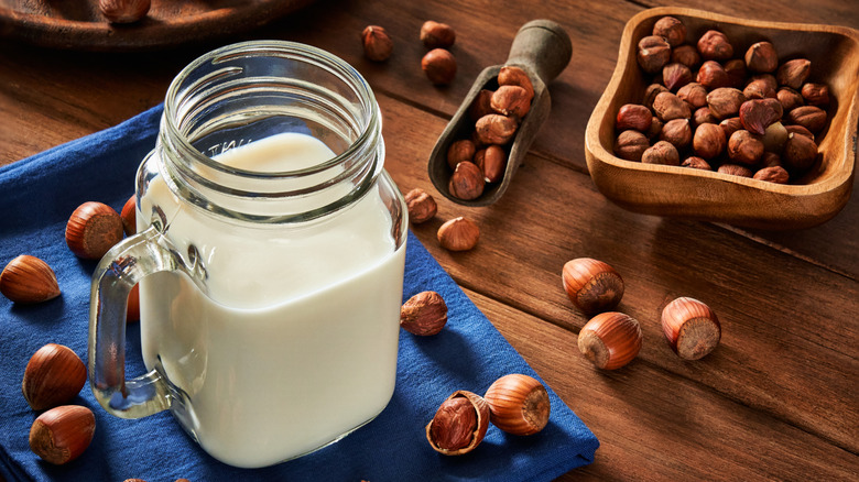 A squared glass of hazelnut milk sitting on a blue cloth, surrounded by scattered nuts on rustic wooden table
