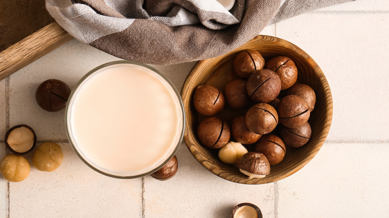 A top-down view of a glass of macadamia milk next to a wooden bowl of macadamia nuts, on a white tile background