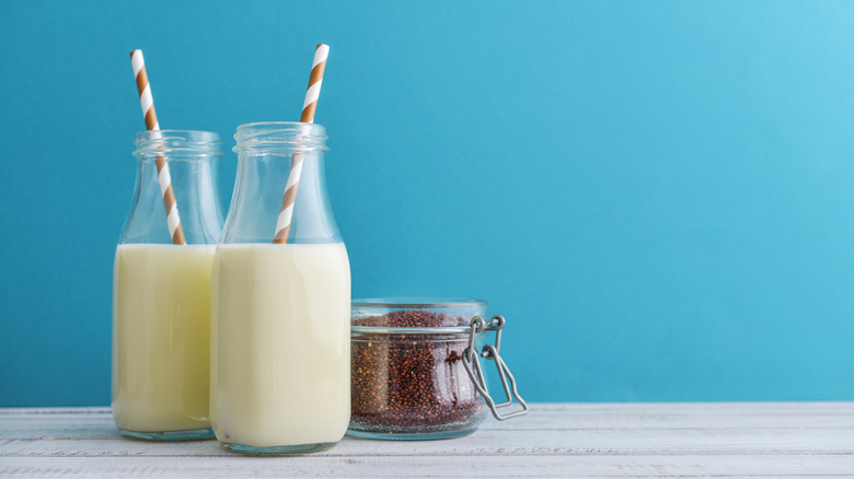 Two bottles of quinoa milk with red and white straws in, next to a closed jar of quinoa seeds on a white table with a blue background