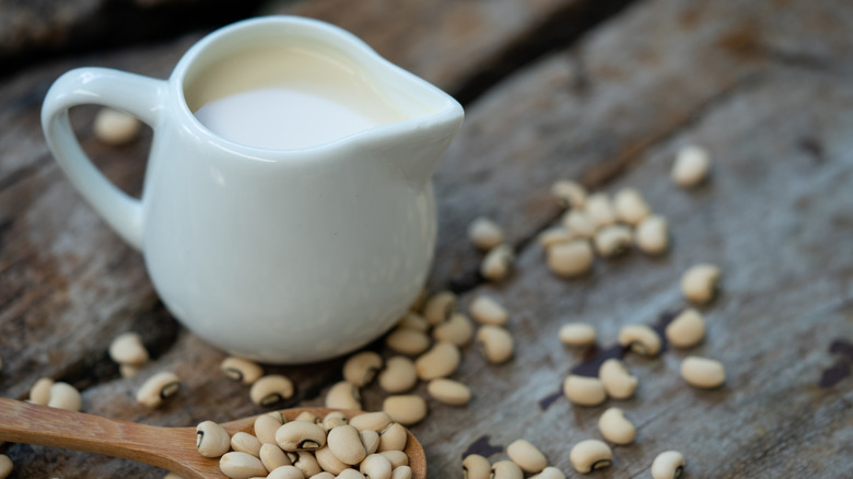 Soy milk in a small white jug next to soy beans on wooden spoon and wooden table.
