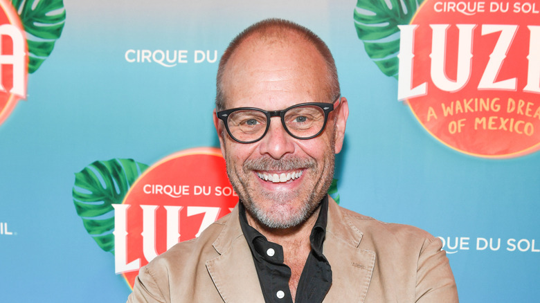 Alton Brown smiling on red carpet while wearing tan jacket.