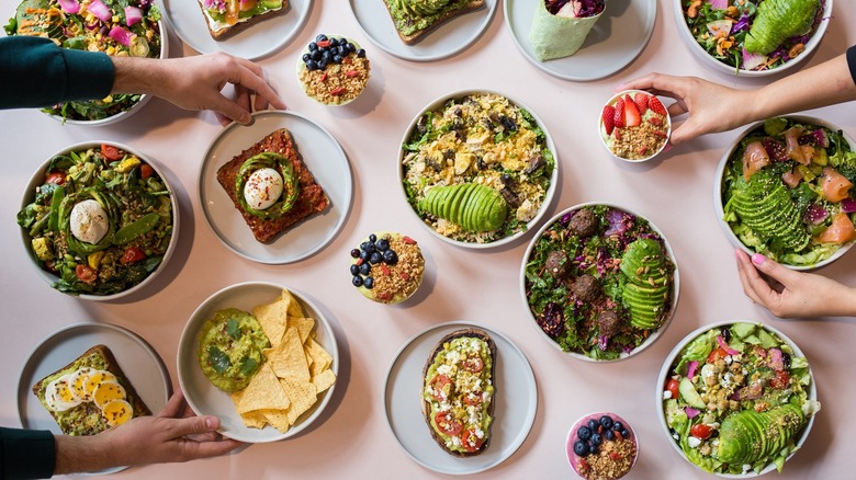 Overhead view of many avocado-based dishes, with hands reaching towards containers