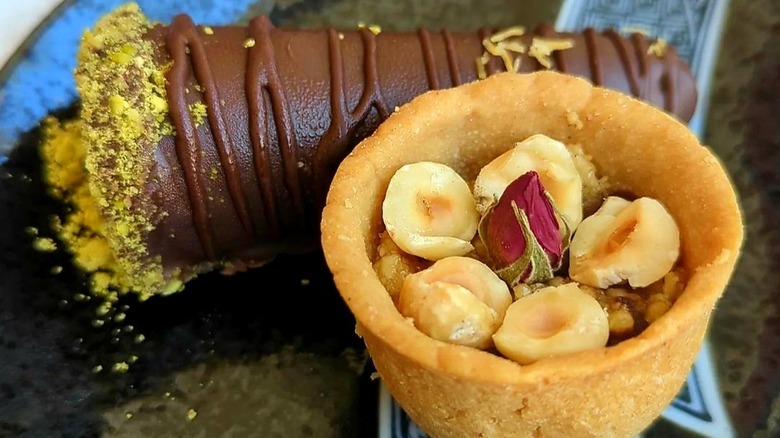 Pastry cup with hazelnuts and rose petals in front of chocolate-dipped pastry with pistachios
