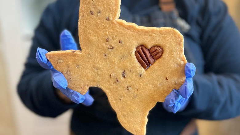 Two gloved hands holding a large pecan shortbread cookie shaped like the state of Texas