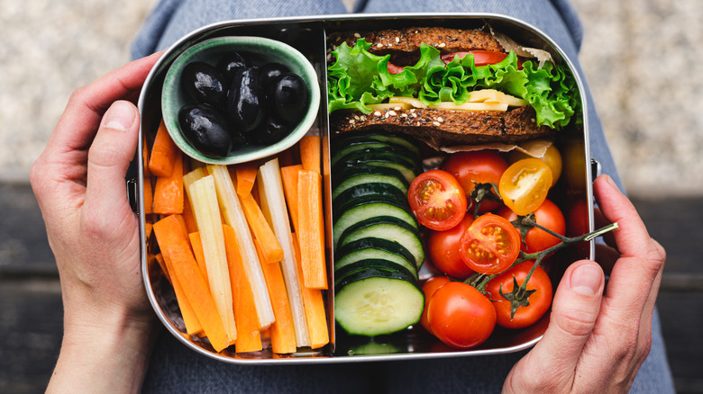 A close up of an adult lunch box with a sandwich and fresh vegetables