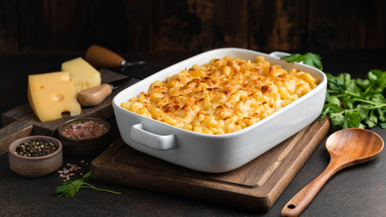 Gourmet macaroni and cheese in a white dish on a wooden cutting board, with a wooden spoon to one side, fancy cheese on a cheese board on the other, and herbs in the background