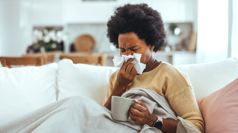Person on couch with mug blows nose into tissue.