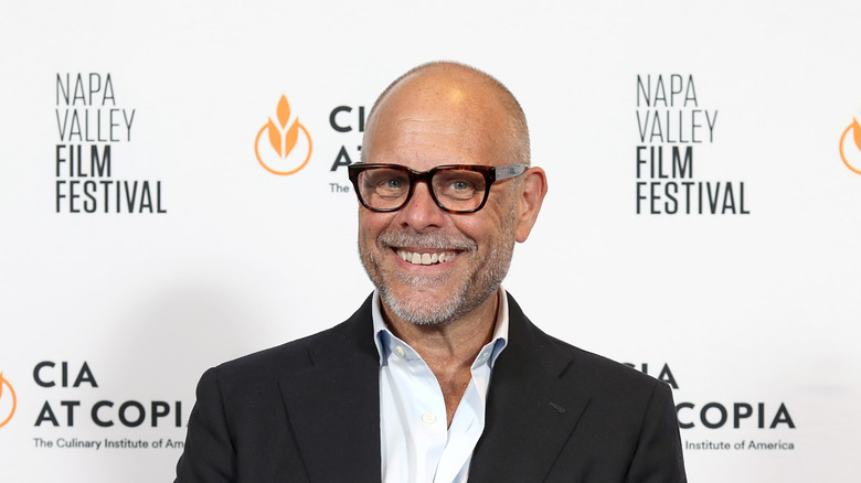 Alton Brown smiling on red carpet, wearing dark jacket.