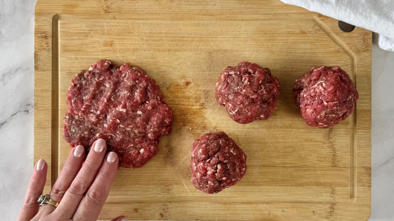 hand touching a raw burger patty on a cutting board with three balls of ground meat