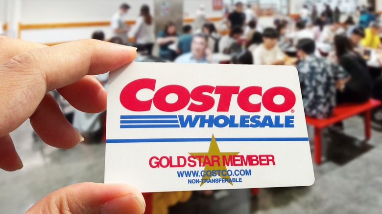 Closeup hand holding Costco card, blurred food court background.