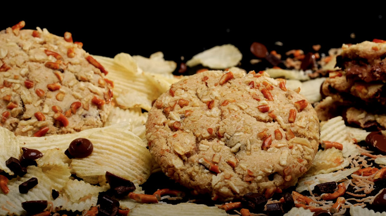 Chocolate Potato Chip Crumbl cookies surrounded by potato chips, pretzels, chocolate chips, and coconut