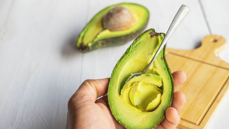 Someone carving out the insides of half an avocado with a spoon over a wooden cutting board, with another half of an avocado with the pit in the background
