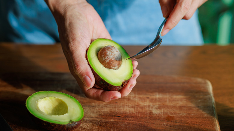 A close-up of someone's hands about to scoop out an avocado's insides with a spoon, with the pit still inside, over a wooden cutting board