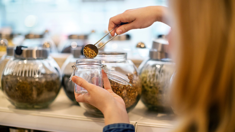 A close-up of a young woman using a metal scoop to fill a glass jar with spices at a no-waste grocery store