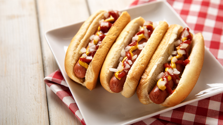 Three hot dogs in bun on white plate over picnic cloth