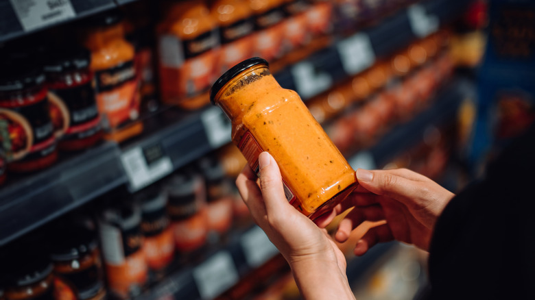 Close-up of person holding jar of sauce in grocery store aisle.