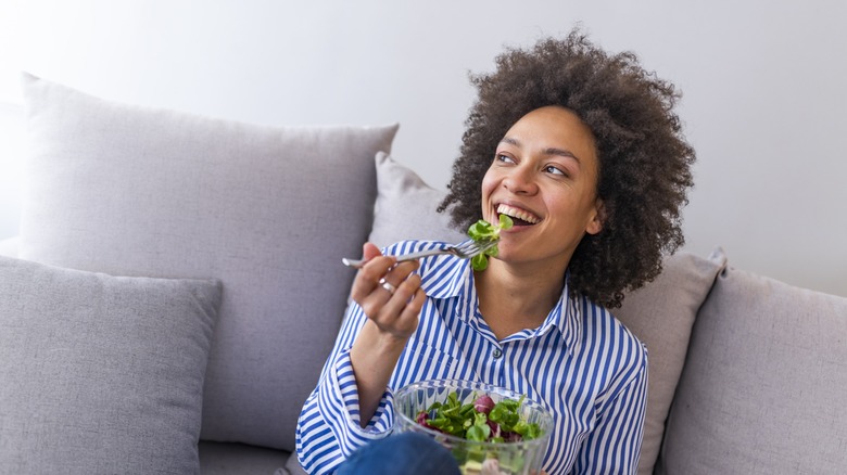 Smiling person eats a green salad while sitting on couch.