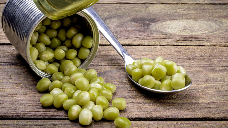 An open can of green peas with some on a silver spoon