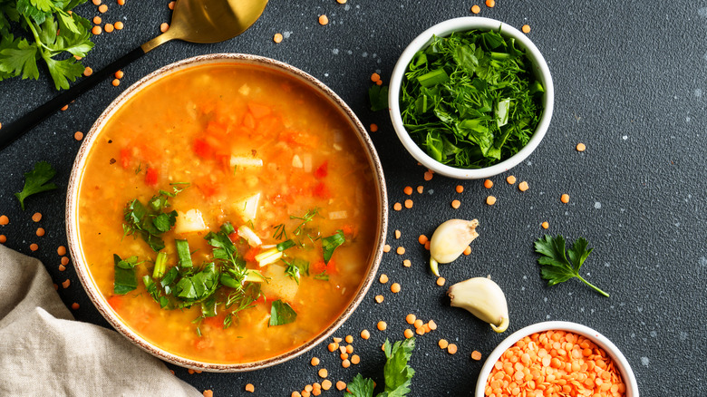 A bowl of lentil soup on a black background, with various other ingredients scattered around