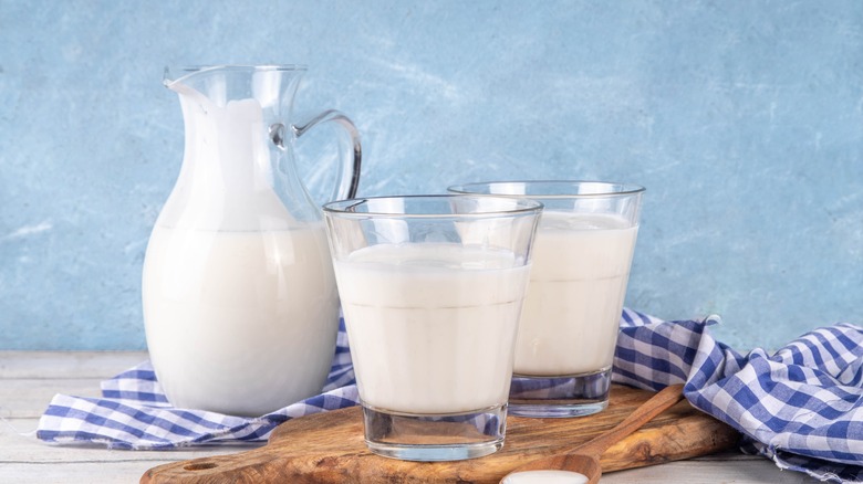 Two glasses and a pitcher of buttermilk on a wooden cutting board and checkered white and blue tablecloth