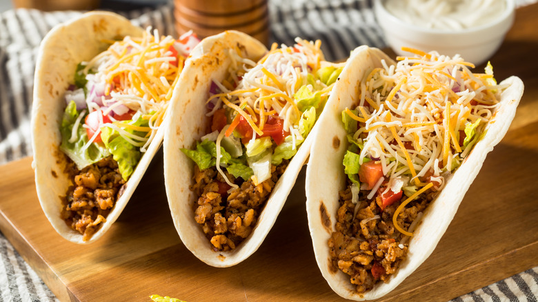 Three flour tortilla tacos with meat, tomato, guacamole, and cheese on wooden cutting board