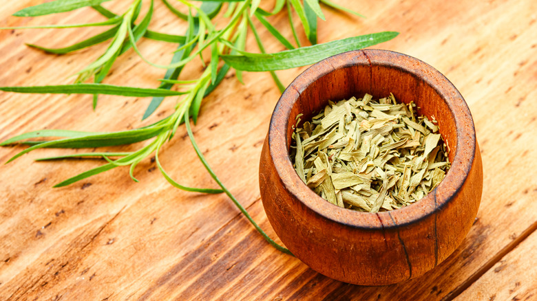 A wooden bowl of dried tarragon next to some fresh leaves